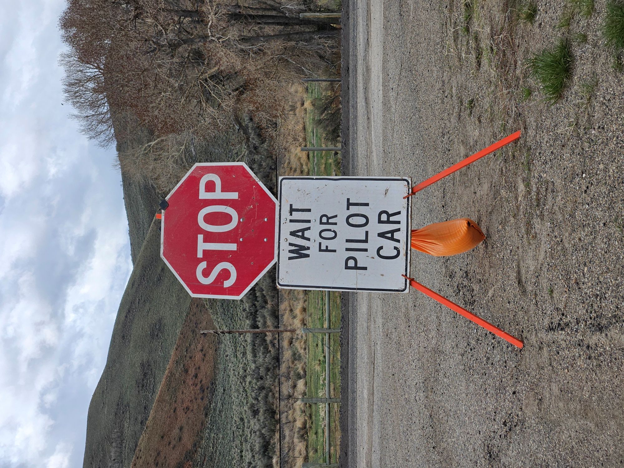 A temporary road construction sign assembly standing on a gravel roadside. A standard red octagonal "STOP" sign is mounted above a white rectangular sign that reads "WAIT FOR PILOT CAR" in black block letters. The signs are supported by an orange metal A-frame stand weighted down by an orange sandbag. The background shows a rural Wyoming landscape with sagebrush-covered hills, a wooden fence, and a cloudy sky near Kemmerer.